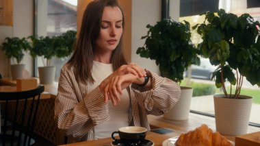 Kafedeki endişeli Kafkasyalı kadın kol saatiyle pencereye bakıyor. Kafeterya masasında bekleyen, omuz silken, sabırsız iş kadını, geç kalan iş görüşmesi.