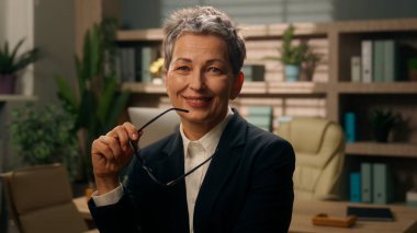 Ofiste düşünceli, yaşlı, düşünceli, beyaz kadın, iş kadını, olgun, orta yaşlı, elinde gözlükleriyle gülümseyen bir işveren, kameraya bakarak bir fikir üretiyor.