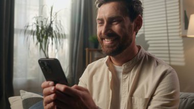 Mutlu adam cep telefonu kullanıyor gülümsüyor Kafkasyalı erkek iş adamı akıllı telefonuna bakıyor iyi haber mesajı neşeli ev sahibi oturma odasındaki kanepede dinleniyor online alışveriş uygulaması teslimatı