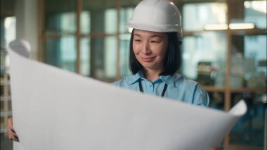 Asyalı kadın mühendis, güvenlik şapkası takıyor, gülümsüyor. Çinli Koreli Japon kız sanayi mimarı müteahhit. Şantiye planı inşaat projesine bakıyor. Elektrik şeması.