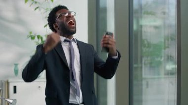 Heyecanlı Afrikalı Amerikalı iş adamı bağırarak ofisteki cep telefonuyla kutlama yapıyor duygusal sevinç içinde etnik iş adamı kazanan erkek şampiyon web akıllı telefon zaferi