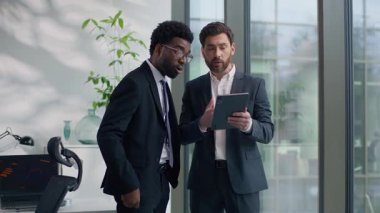 İki farklı etnik çeşitlilikteki iş adamı, dijital tablet bilgisayardan bakarak online başarıyı tartışıyor. İyi haber, çok ırklı iş adamları ofiste yeni başlayan büyümeden bahsediyorlar.