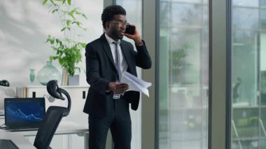 Afro-Amerikalı etnik iş adamı cep telefonu sorunu yüzünden tartışıyor ofiste kağıtları tutuyor öfkeli iş adamı akıllı telefonla bağırıyor ve belgeleri pencereden dışarı atıyor.