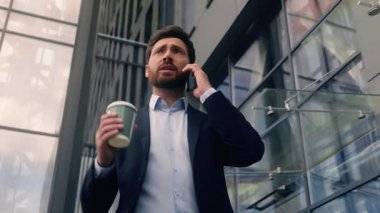 Orta yaşlı, Kafkasyalı, ciddi iş adamının elinde bir fincan kahve ile şehirde cep telefonuyla konuşmasından bıkmış, yürüyen iş adamı, ofis binasının yakınında yürüyor, akıllı telefon ile iletişim kuruyor.
