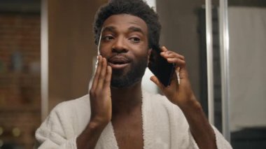 Aynada yansıyan banyoya bakan Afrikalı bir adam cep telefonuyla konuşan, yüz bakımına dokunan, sakallı, sakallı, erkek, otel banyosunda akıllı telefon görüşmesi yapan, sakallı, sakallı bir adam.