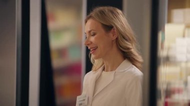 Sağlık çalışanı kadın eczacı eczanesinde konuşuyor. Eczanede tıbbi yardım alıyor. Müşteri ilaç paketini kasiyerden alıyor.