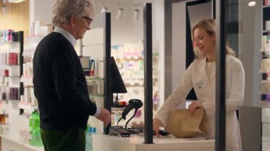 Eczacı kadın eczacının tavsiyesine göre ilaç satın alan yaşlı bir adam ilaç paketini kasiyer gişesinden, eczane tıbbi yardım ilaç vitaminlerinden satın alır.