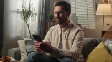 Gülümseyen adam zımbırtısı bağımlısı iş adamı Kafkasyalı ev sahibi kanepe oturma odasında akıllı telefon kullanarak rahatlıyor mutlu erkek cep telefonuyla internetten alışveriş yapıyor sosyal medya alışverişinde gülüyor mesajın tadını çıkarın