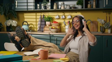 Latin Amerikalı etnik kadın mutlu genç kız lise öğrencisi rahatlamış kız öğrenci öğrenciliği yapıyor ev mutfağında cep telefonuyla şarkı seçiyor kulaklıkla müzik dinliyor, müzik dinliyor, dans ediyor.