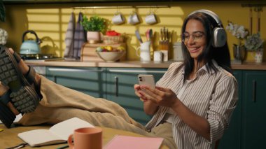 Latin Amerikalı etnik kadın mutlu genç kız lise öğrencisi rahatlamış kız öğrenci öğrenciliği yapıyor ev mutfağında cep telefonuyla şarkı seçiyor kulaklıkla müzik dinliyor, müzik dinliyor, dans ediyor.