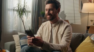 Gülümseyen adam zımbırtısı bağımlısı iş adamı Kafkasyalı ev sahibi kanepe oturma odasında akıllı telefon kullanarak rahatlıyor mutlu erkek cep telefonuyla internetten alışveriş yapıyor sosyal medya alışverişinde gülüyor mesajın tadını çıkarın