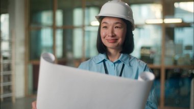 Asyalı kadın mühendis, güvenlik şapkası takıyor, gülümsüyor. Çinli Koreli Japon kız sanayi mimarı müteahhit. Şantiye planı inşaat projesine bakıyor. Elektrik şeması.
