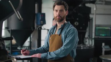 Kafkasyalı bir adam fabrikada çalışıyor kahve çekirdekleri kavrulmuş kağıt not yazıyor kavrulma makinası denetleme teknolojisi erkek adam önlüklü adam kameraya bakıyor profesyonel baristaya bakıyor.