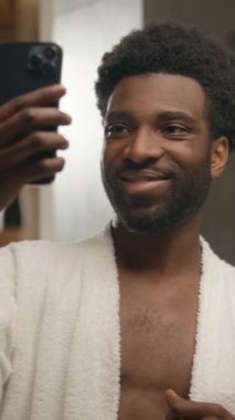 Afro-Amerikalı blogger manken mankeni selfie fotoğraflı cep telefonu banyoda cilt bakımı blogu sosyal medya yakışıklı gülümseyen etnik erkek adam ev banyosunda kozmetik video akıllı telefon pozu veriyor.