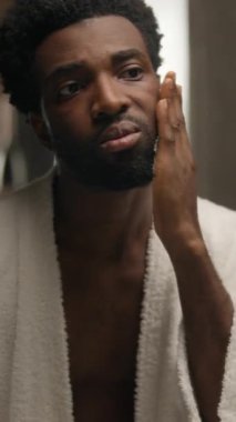 Afro-Amerikalı bir adam banyoda köpüklü jöle sürüyor sakal traş ediyor sakal bakımı erkek lekeli erkek makyaj köpüğü güzellik ürünü tıraş olurken aynadaki yansımaya bakıyor traş edilmemiş üzgün adam elma kremi kozmetik ürünleri.