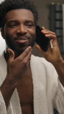 Banyodaki Afrikalı Amerikalı adam sabah duştan sonra cep telefonuyla konuşuyor sakalını kesiyor gülümseyen sakallı adama dokunuyor arkadaşça sohbet ediyor otel banyosunda cep telefonuyla iletişim kuruyor.