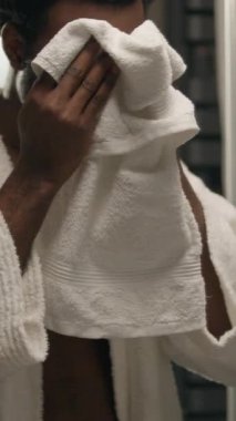 Afro-Amerikalı bir adam banyoda yüzünü yıkıyor. Havluyla yüzünü siliyor. Cilt bakımını tazeliyor.