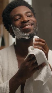 Afrikalı Amerikalı adam sakalını tıraş ediyor. Banyo sakallı adam. Havluyla köpük siliyor.