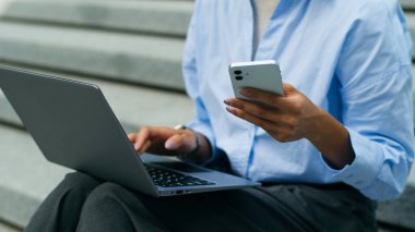 Tanımlanamayan Afro-Amerikan öğrenci kız iş kadını kadın işkadını. Şehir dışında oturan iş kadını. Bilgisayarda cep telefonu akıllı telefonları kullanarak bilgisayar yazıyor.