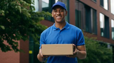 Gülümseyen Afro-Amerikan etnik kurye portresi, şehirde durup gülümsüyor, kameraya bakıyor, postacı, postacı, postacı, mavi üniformayla teslimat yapıyor.