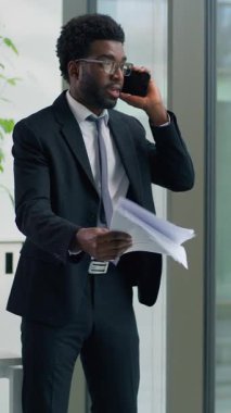 Dikey bakış Afrikalı Amerikalı etnik iş adamı kızgın cep telefonu problemini tartışırken ofiste kağıtları tutuyor öfkeli iş adamı bağırıyor akıllı telefondan belgeler atıyor pencereden dışarı bakıyor.