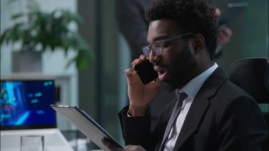 Endişeli Afrikalı Amerikalı işadamı, cep telefonuyla konuşan, iş sözleşmesi belgelerini okuyan, iş sözleşmesi belgelerini okuyan, ofis raporunda sorun çıkaran bir müşteriyle görüşüyor.