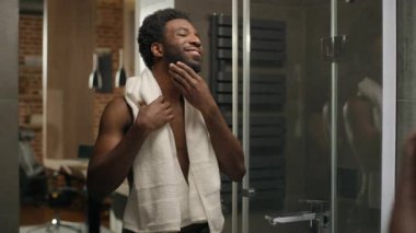 Afro-Amerikalı çıplak adam, aynada gülümseyen sakallı erkek adam, sabah tıraş oluyor, duş alıyor, yüzünü tıraş ediyor, cildini tıraş ediyor, makyaj yapıyor, losyon kremini nemlendiriyor.