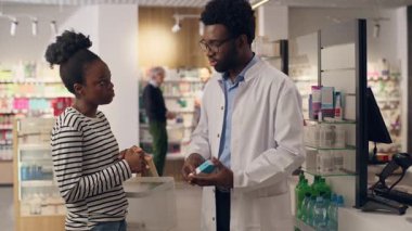 Afro-Amerikalı erkek eczacı, eczanedeki müşterilere, ilaç dozajı semptomlarını tavsiye etmede yardım ediyor. Kadın, eczanedeki sağlık hizmetlerinde vitamin satın alıyor. Laboratuvar önlüğü tıbbında danışmanlık satıyor.