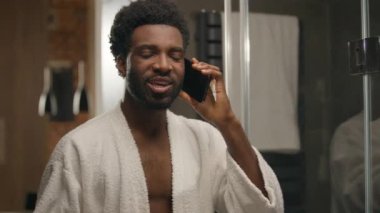 Bornozlu Afrikalı Amerikalı bir adam cep telefonuyla konuşuyor aynaya bakıyor gülümseyen bir etnik adam vücut bakımı sabah duş hijyenik sakallı bir erkek otel banyosunda akıllı telefon konuşması yapıyor.