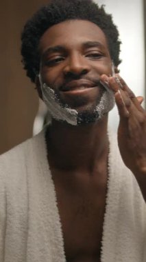 Banyoda sakal tıraş eden Afrikalı Amerikalı adam köpüklü jel cilt bakımı uyguluyor sakallı erkek tıraş köpüğü tıraş edilmemiş erkek yüzüne bakıyor pürüzsüz krem kozmetik güzellik uygula