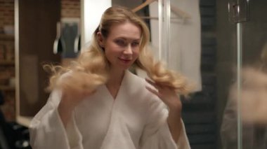 Tarz sahibi, güler yüzlü kadın bornozu saç stili saç stili kozmetik banyo Kafkasyalı neşeli kız banyo duşunda muhteşem sarışın kadın manken aynaya bakıp kuaför güzelliğine dokunuyor.