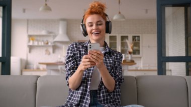 Mutlu gülücükler, mutlu dans, tatmin edici özgür gülümseme, Kafkasyalı kadın bayan ev sahibi, cep telefonu ses uygulaması, kulaklıkla müzik, evde şarkı sesi.