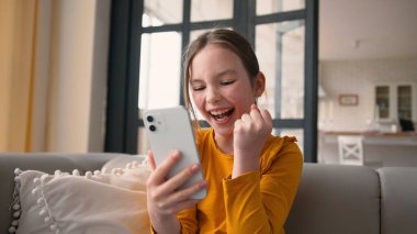 Neşeli küçük beyaz kız elinde telefon cihazı tutarken gülümseyen kız seviniyor. Mobil oyundaki zaferi kutlayan liseli kız çevrimiçi uygulama çığlığı harika sonuç evde iyi haber