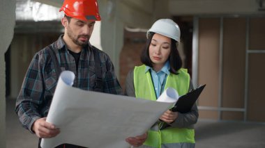 Üniformalı çok ırklı müteahhitler taslak teftişi iş mühendisleri Kafkasyalı erkek Koreli kadın inşaat projesini tartışıyorlar.