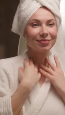 Güzel, mutlu, beyaz bir kadın bornoz havlu masajı yapıyor boyun kremi sürüyor, gülümsüyor, genç, güzel bir bayan model aynaya bakıyor yumuşak tene dokunuyor duşun keyfini çıkarıyor, rutin losyon banyosu yapıyor.