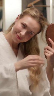 Güzel, mutlu, genç bir kız saçını tarıyor banyoda görünüş alıyor beyaz kadın bornozu cilt bakımının keyfini çıkarıyor evde banyo banyosunda saç fırçası hazırlıyor.