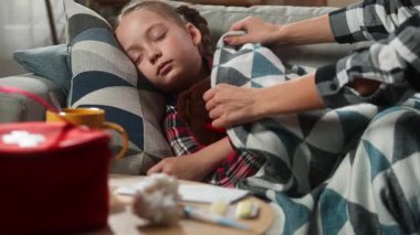 Beyaz aile, ev hekimliği, sağlık sorunları, uyuyan çocuk, hasta kız, kanepe üzerinde uyukluyor, şefkatli anne, battaniyeli küçük kız, alnına dokunuyor, ateşi kontrol ediyor.