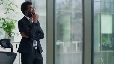 Sorunu çözmeyi düşünen Afro-Amerikalı işadamının ofis penceresine bakması sorunlu iş başarısızlığına karar vermesi yorgun iş adamı zor karar vermek cama yaslanmak erkeğin tereddüt ettiğini düşünüyor.