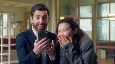 Ofiste cep telefonu başarısını kullanan Koreli beyaz iş kadını heyecanla iş arkadaşı ortaklarına online zafer zaferi için tezahürat yapan akıllı telefon görüntüsü sunuyor.