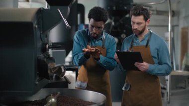 Çok yönlü iki erkek kahve fabrikası çalışanı fasulye kızartma makinası kalite kontrol listesi klasörü Kafkasyalı Afrikalı uzman kahve üretimi endüstriyel malzeme teftişi içeceğini tartışıyor.