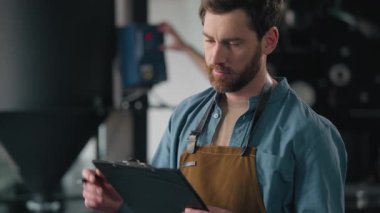 Erkek işçi Kafkasyalı teknisyen kahve kızartma fabrikası kontrol makinesini denetliyor kontrol listesi kalite kontrol kafein imalatçısı şirket sahibi üretim endüstrisine notlar yazıyor