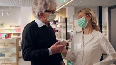 Eczane sağlık danışmanlığındaki yaşlı adam. Maskeli kadın eczacı. Antiviral haplar öneriyor.