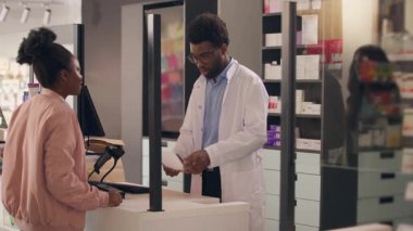Afro-Amerikalı erkek eczacı, eczane reçeteli ilaç müşterisine yardımcı oluyor. Kadın, ilaç alıyor. İlaç alıyor, vitamin alıyor, ilaç alıyor.