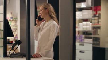 Eczane kasiyerindeki beyaz kadın cevap veriyor. Cep telefonuyla konuşuyor. İnternetten ilaç siparişi veriyor.
