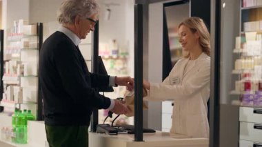 Eczane profesyonel tıbbi yardım gülümseyerek kadın eczacı danışmanı. Kasiyerde ilaç satan yaşlı adam. Kağıt torbada ilaç alan yaşlı bir müşteri.