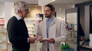 Olgun, yaşlı bir eczacı eczaneden ilaç almaya yardım ediyor. Kalp hapları, sağlıklı vitaminler, güler yüzlü erkekler. Müşterinin sağlık hizmetlerine yardım eden doktor.