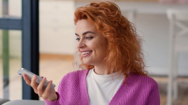 Mutlu, güler yüzlü, beyaz kadın. Evdeki kanepe koltuğunda oturan, cep telefonu uygulaması kullanan, gülümseyen, sesli mesaj kaydeden, akıllı telefonlu akıllı telefon konuşmalarını kaydeden.