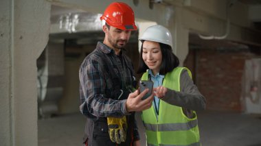 İki çok ırklı inşaatçı cep telefonu tasarım uygulaması kullanarak Japon kadını beyaz erkek olarak gösteriyorlar. Hardhat mühendisleri iş müfettişleri akıllı telefon yeşil ekranına göz atıyorlar.