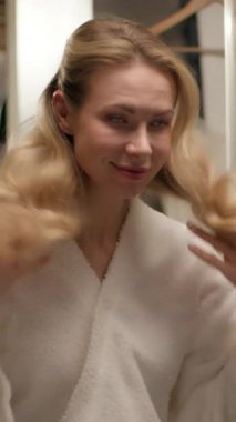 Tarz sahibi, güler yüzlü kadın bornozu saç stili saç stili kozmetik banyo Kafkasyalı neşeli kız banyo duşunda muhteşem sarışın kadın manken aynaya bakıp kuaför güzelliğine dokunuyor.