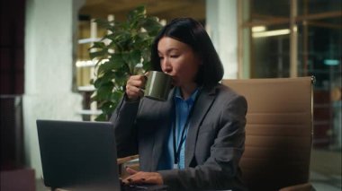 Pozitif Asyalı Koreli Çinli iş kadını dizüstü bilgisayar yazan kız elinde fincanla kahve içiyor ofis çalışanı yönetici yönetici kadın içeride çalışıyor.